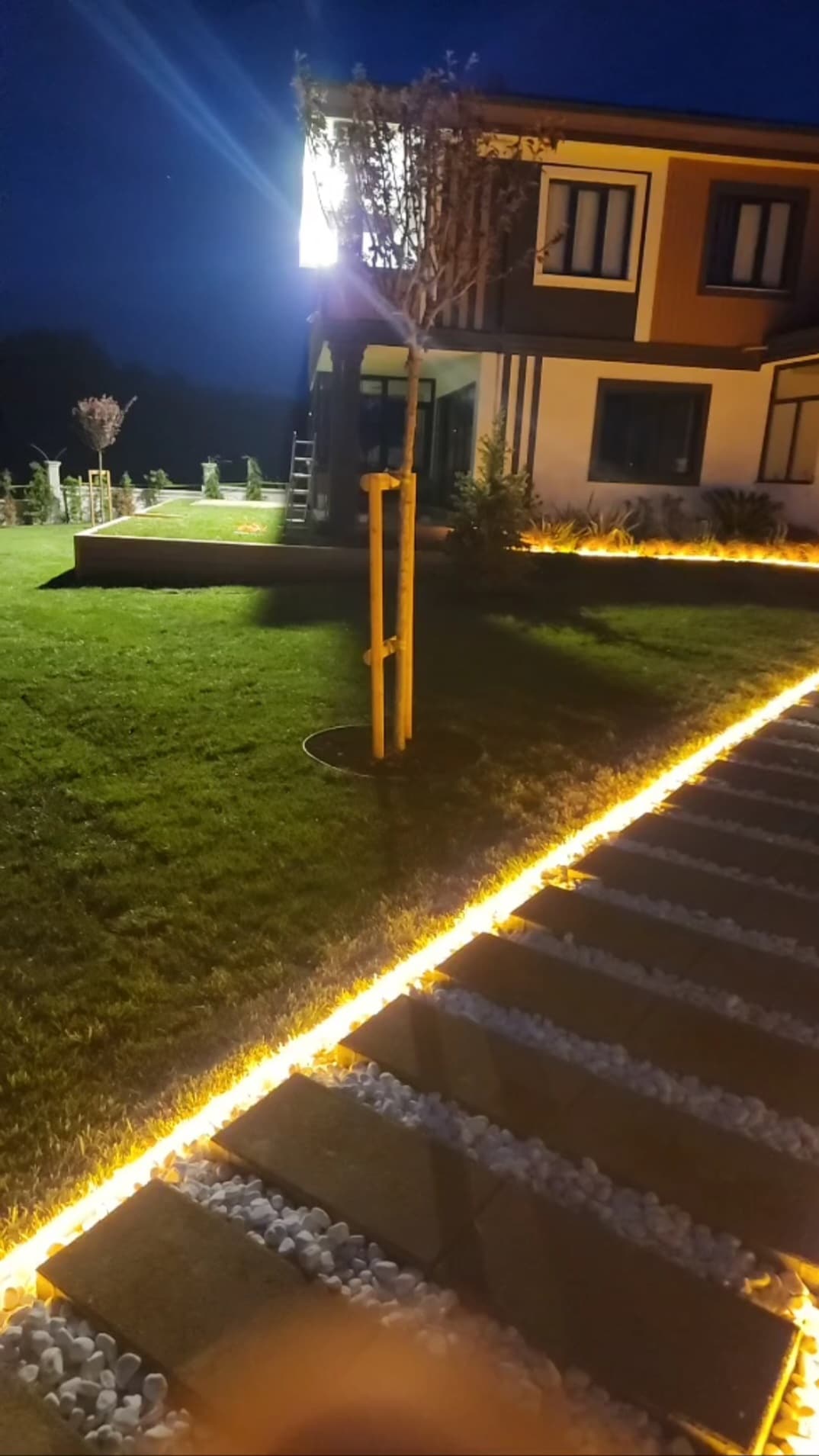 The exterior of the property is highlighted at night, showcasing a well-manicured lawn and a pathway illuminated by soft, warm lighting. Decorative stones border the path, leading to the house, which features large windows reflecting the outdoor lighting for a welcoming appearance.