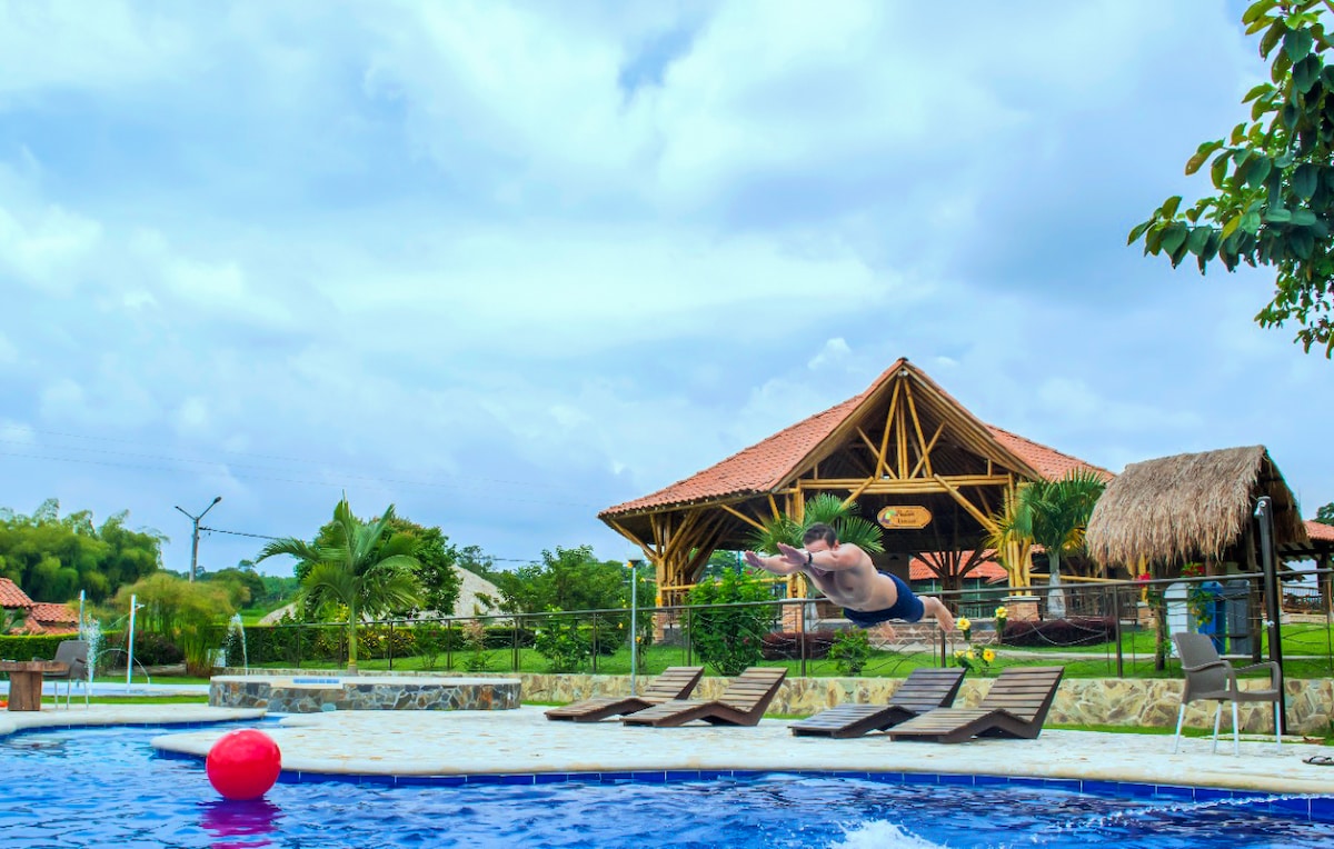 A swimmer is captured mid-air above a blue pool, with a colorful red beach ball floating nearby. In the background, a thatched-roof structure is surrounded by lush greenery, and several lounge chairs are neatly arranged by the poolside.