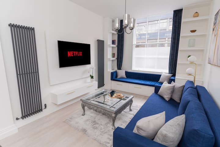 Модерен простор за одмор со отворен план со удобен кауч на спуштање, HD телевизор со рамен екран, мек сив килим и масичка за кафе.