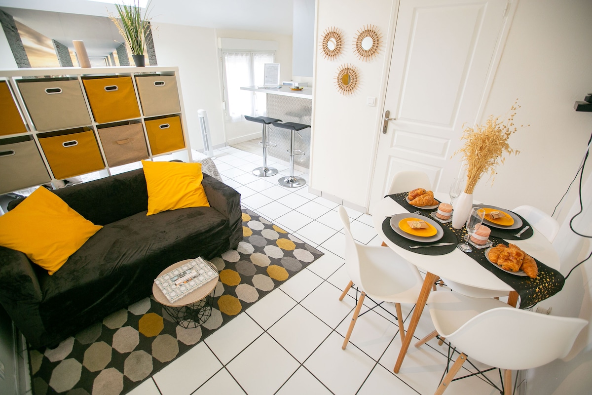 A welcoming living area features a dark sofa adorned with bright yellow pillows, set against a patterned rug. A dining table with four white chairs is prepared with plates and pastries, while a modern bar area with stools is visible in the background.