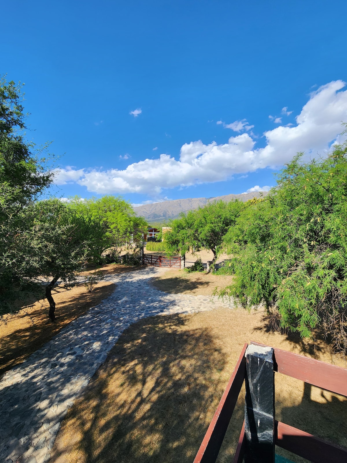 A view of the property is offered, featuring a pathway lined with trees leading towards the entrance. Distant mountains are visible under a clear blue sky with scattered clouds, creating a serene outdoor environment.