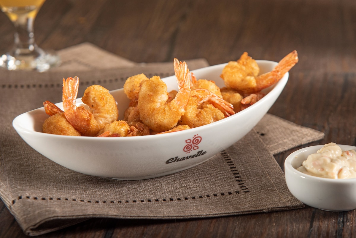 A white ceramic dish presents a generous serving of golden-brown fried shrimp, garnished with tails. Next to the dish, a small white bowl contains creamy dipping sauce. The setting features a textured brown tablecloth, enhancing the visual contrast.