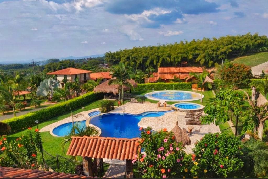 A scenic view of the hotel grounds showcases multiple outdoor swimming pools, surrounded by tropical plants and vibrant flowers. Thatched-roof seating areas are visible, offering shaded spots for relaxation. Lush greenery and hills provide a picturesque backdrop under a partly cloudy sky.