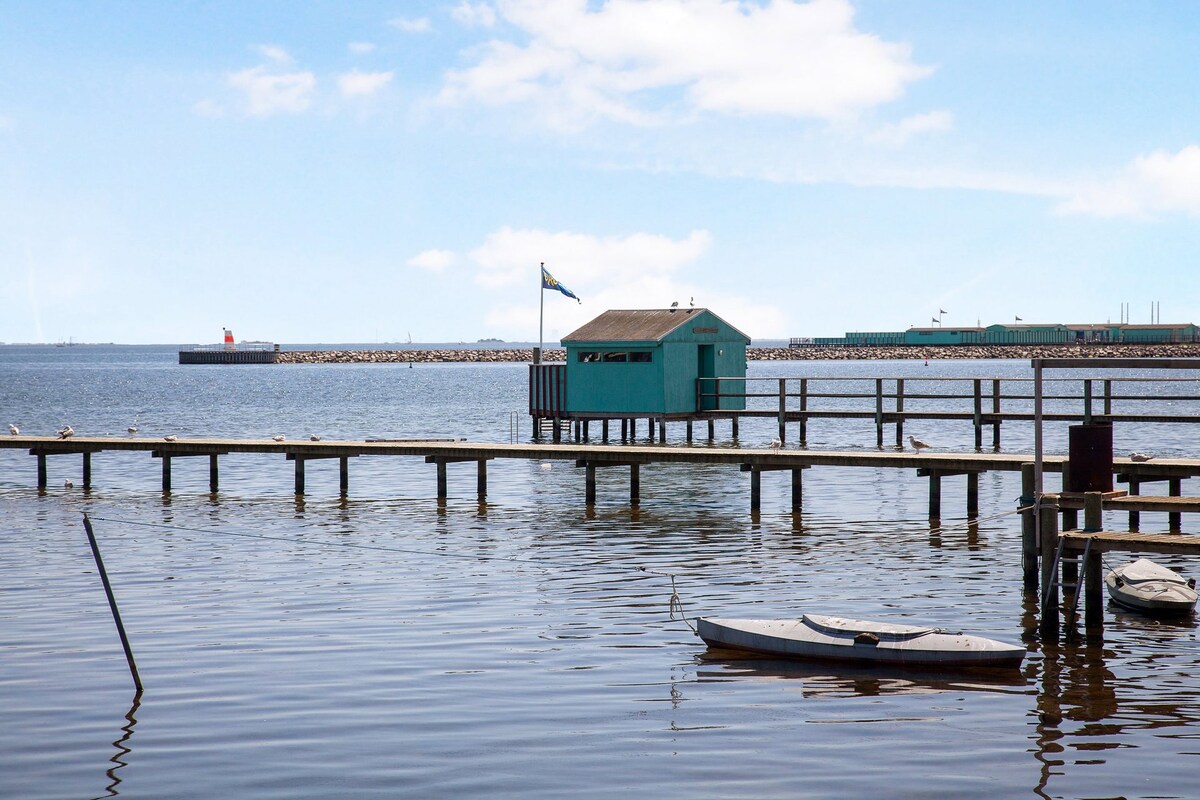 A wooden dock extends over still water, leading to a quaint blue structure with a flag. The reflection of the building can be seen in the water, along with several boats nearby. In the background, a lighthouse and a cargo ship are visible along the shoreline.
