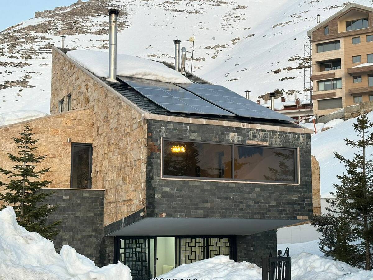 A modern mountain retreat is showcased, featuring a stone and dark-hued exterior with large windows that reflect the surrounding snowy landscape. Solar panels are installed on the roof, highlighting eco-friendly features. Snow blankets the ground, creating a serene winter atmosphere.
