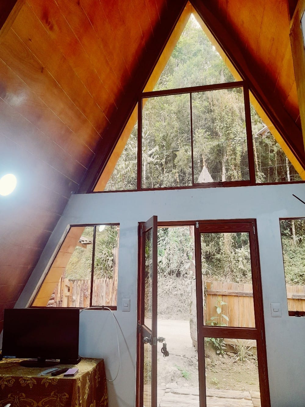 A triangular cabin structure features large glass windows that frame views of lush greenery. Natural wood and white walls create a bright ambience. A small television is positioned on a table, with a doorway leading to an outdoor pathway, inviting in natural light.