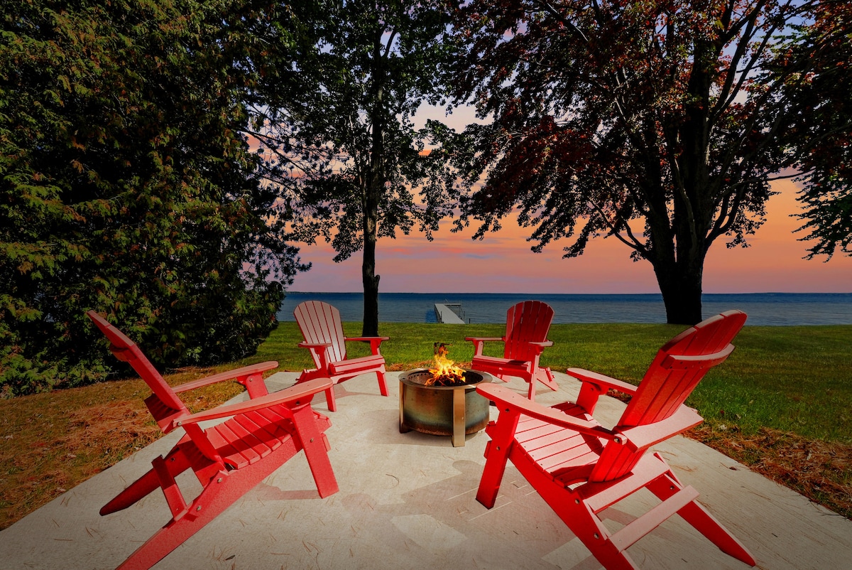 A cozy fire pit area is surrounded by five red Adirondack chairs, positioned on a stone patio. Trees line the background, with a view of the lake and colorful sunset illuminating the sky.