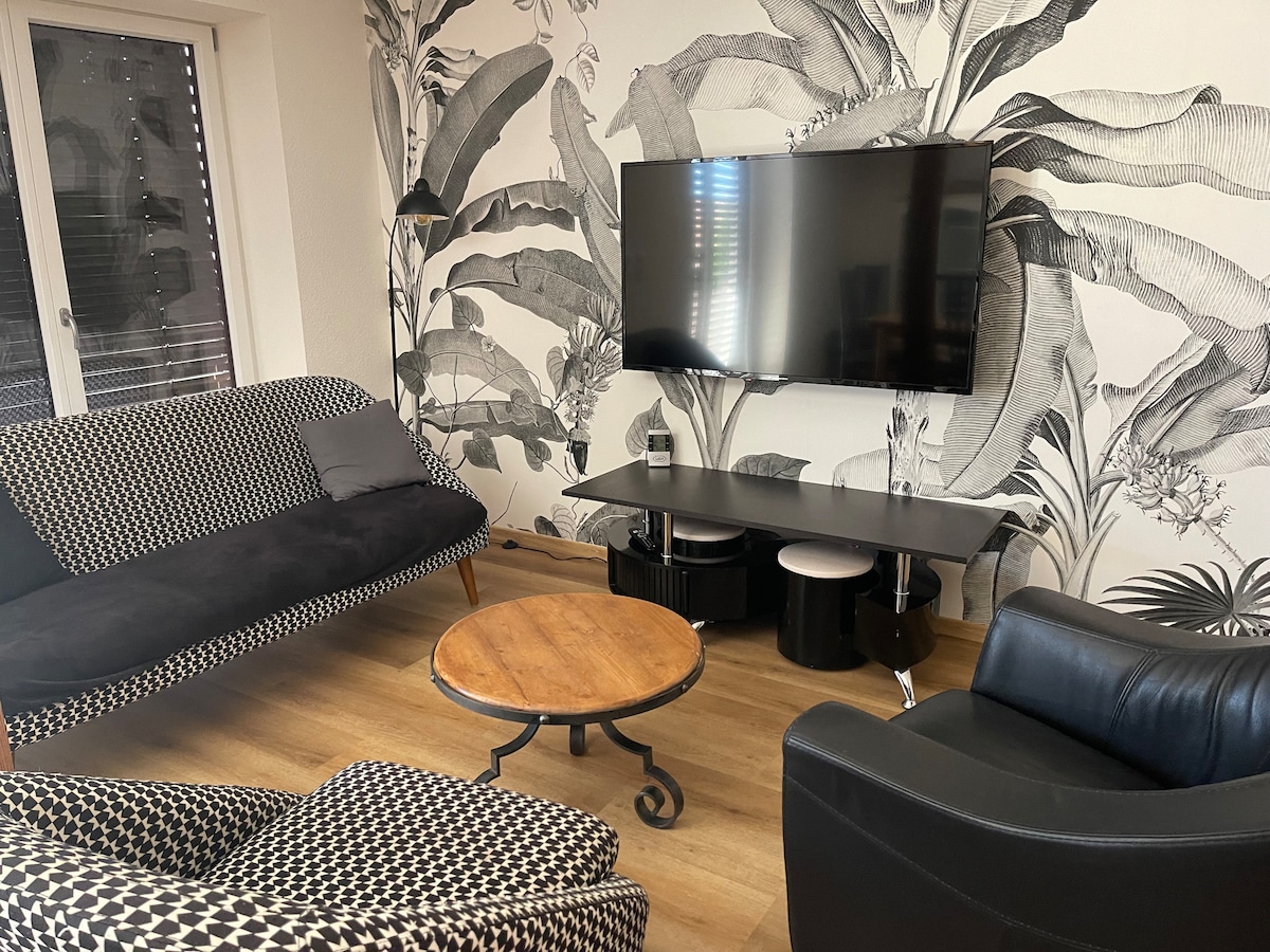 A modern living room is presented with bold botanical wallpaper. A flat-screen TV is mounted on the wall above a sleek black media console. Two stylish sofas face a circular wooden coffee table, while natural light filters through the windows adorned with minimalistic blinds.