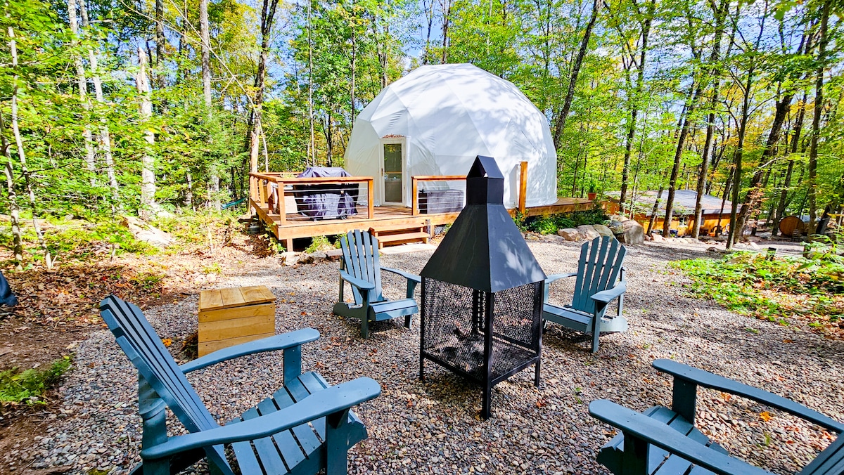 A geodesic dome is nestled among trees, featuring a spacious deck for outdoor enjoyment. Surrounding a central fire pit, several adirondack chairs invite relaxation, while a hot tub can be seen on the deck, enhancing the nature-inspired setting.