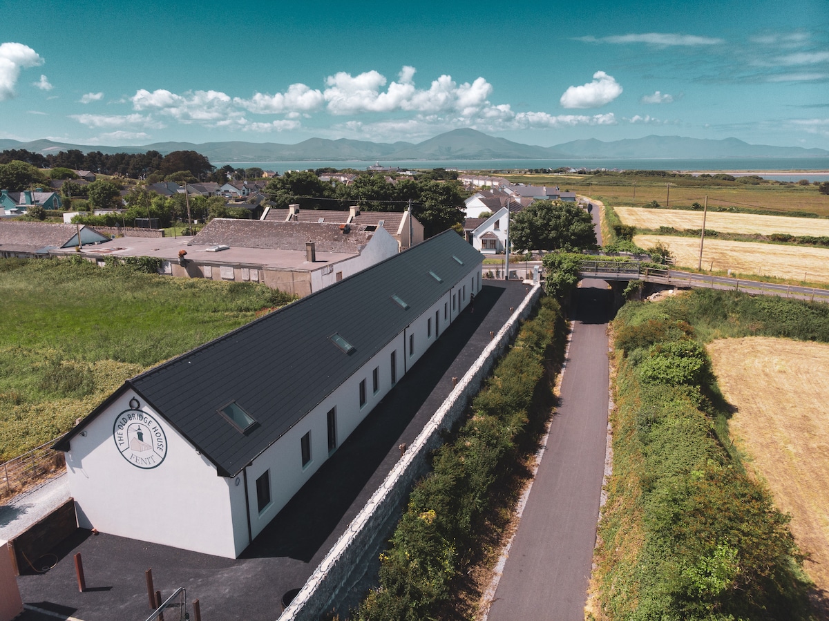 An aerial view displays the sleek, modern design of the Old Bridge House Cottages, set against a backdrop of greenery and distant mountains. A straight pathway leads toward the property, flanked by natural landscapes and residential buildings.