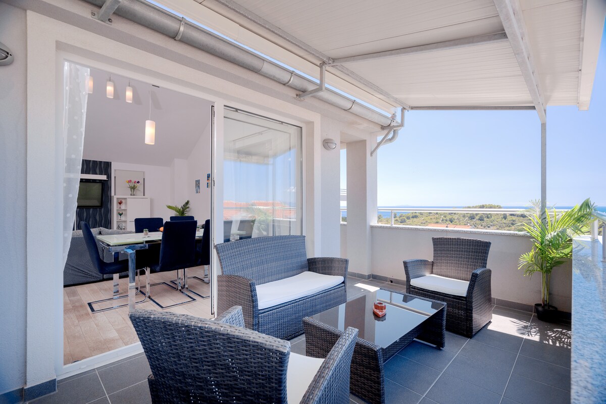 A serene outdoor seating area features wicker armchairs and a glass-topped table, inviting relaxation. The balcony provides unobstructed views of the sea and surrounding landscape through expansive sliding glass doors, ensuring a seamless connection to the indoor living space.