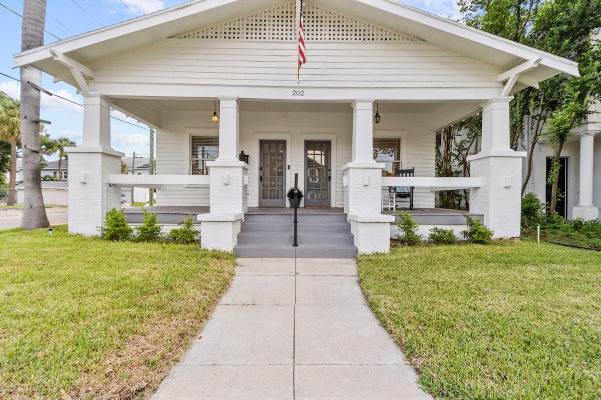 A charming white house with a large front porch is framed by well-maintained greenery. Two front doors are flanked by evenly spaced porch lights. An American flag is displayed above the entrance, adding a welcoming touch to the exterior.