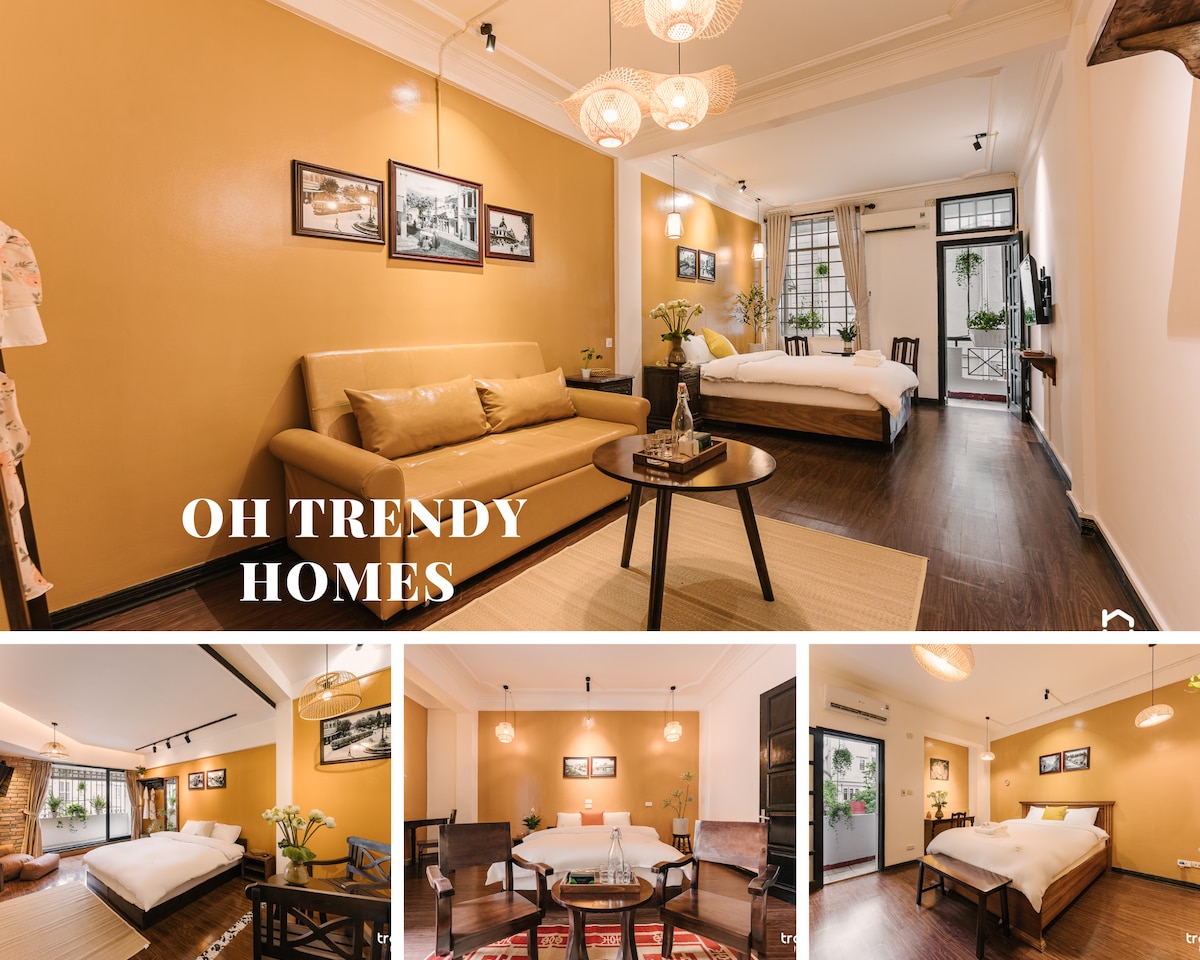 A cozy living space is presented with a beige wall and wooden flooring. A light brown sofa faces a round coffee table, complemented by floral centerpieces. Nearby, two beds with white linens are arranged, and framed photographs add a touch of decor. Natural light enters through multiple windows.