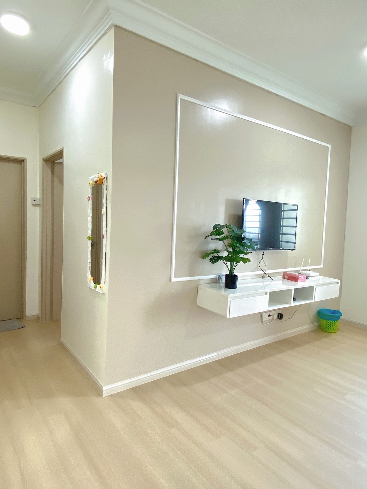 A modern living area includes a wall-mounted television above a sleek white console. A small potted plant adds a touch of greenery. The walls are painted in a soft beige, with subtle decorative molding, and a mirror is positioned on the left side, reflecting light into the space.