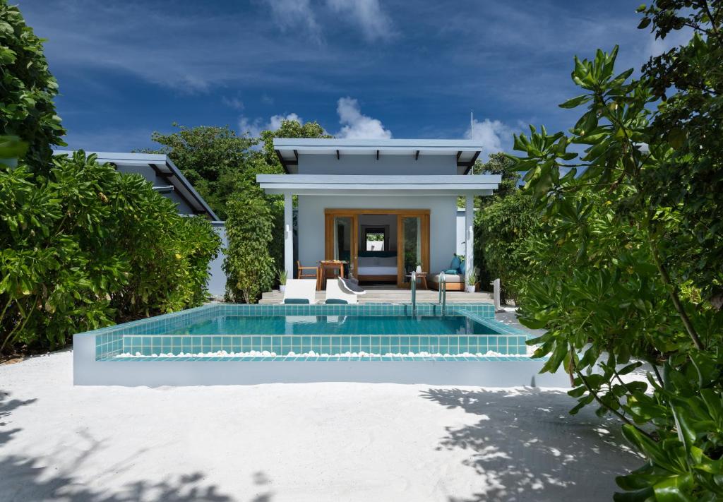 A beach villa is surrounded by lush greenery, featuring a private pool with turquoise tiles. The white sandy area invites relaxation, while the modern structure includes large windows and a welcoming entrance. Clear blue skies provide a bright backdrop, enhancing the serene atmosphere.