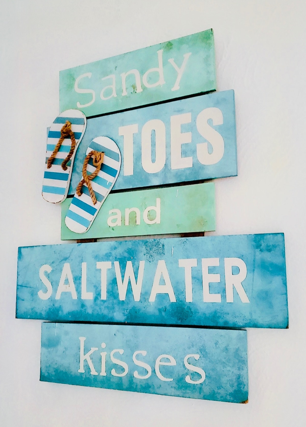 A decorative wall display features layered wooden signs, each painted in shades of blue and green. The phrases 'Sandy Toes' and 'Saltwater Kisses' are prominently displayed in white lettering. A pair of flip-flops is attached, adding a playful coastal element to the decor.