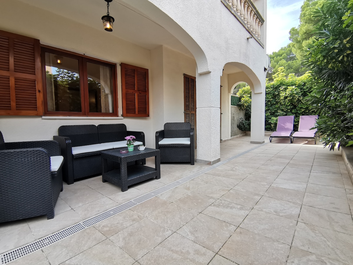 A terrace area is featured with two loungers and a seating arrangement that includes a table and four chairs. Lush greenery surrounds the space, providing a sense of privacy. Sunlight filters through the windows, and the floor is covered in neutral-toned tiles.
