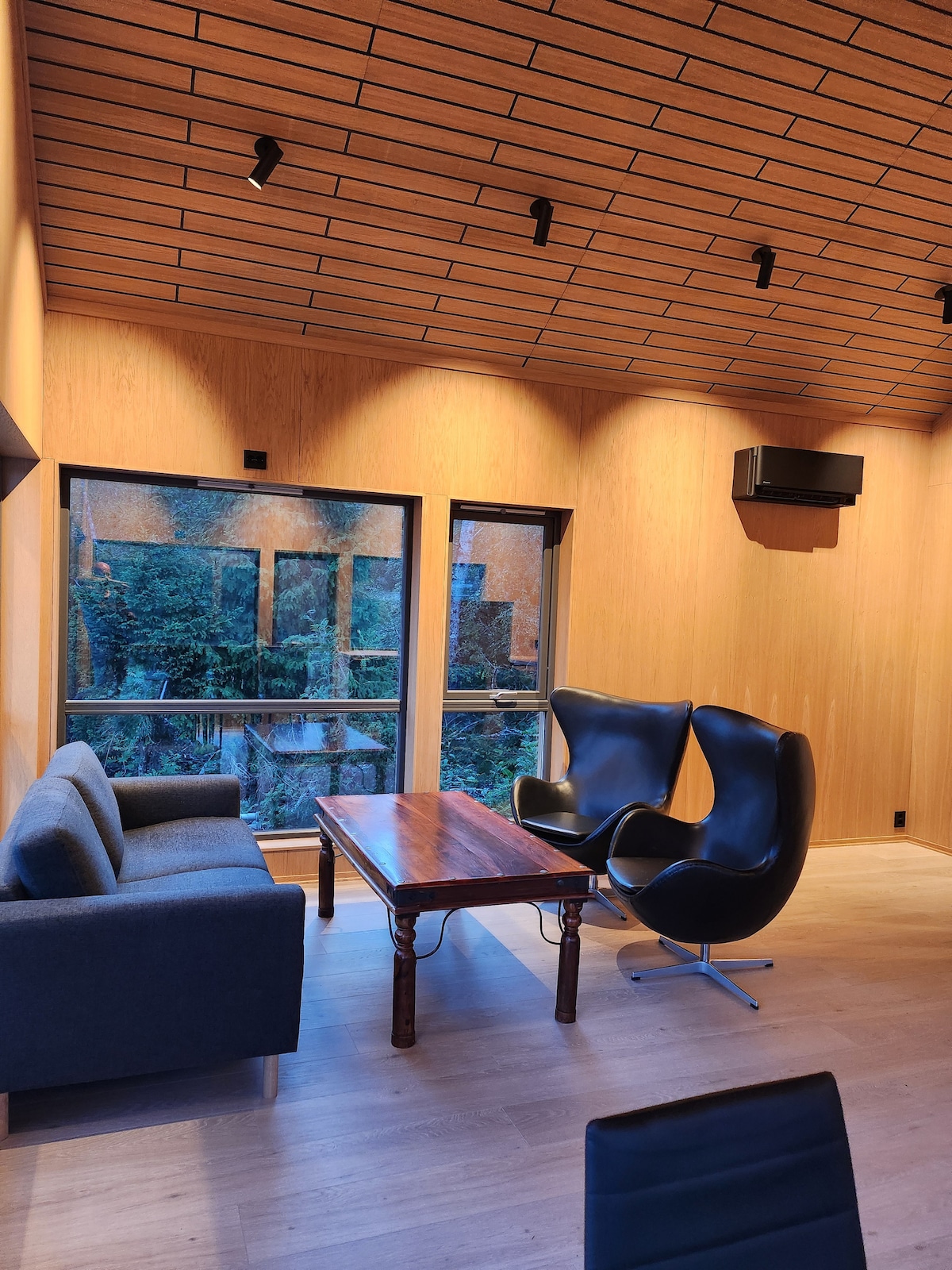A modern living area is highlighted by large windows offering views of the surrounding greenery. It features a gray sofa, a classic wooden coffee table, and two sleek black chairs that invite relaxation. Warm wooden walls complement the stylish lighting above.