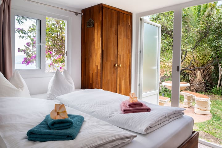 Das Zimmer Mango Süd liegt direkt am Garten und bietet einen wunderschönen Meer- und Gartenblick. Es ist mit stillvollen Holzmöbeln ausgestattet, darunter ein Schrank und ein Schreibtisch und ein Doppelbett, gemütlich und ideal zum entspannen.