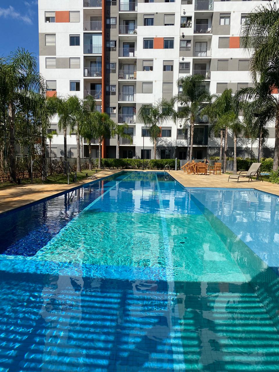 A large swimming pool is framed by palm trees and lounge chairs, reflecting the building's modern architecture. The pool area features a spacious deck and is designed for both relaxation and leisure, with clear blue water inviting guests to enjoy a refreshing swim.