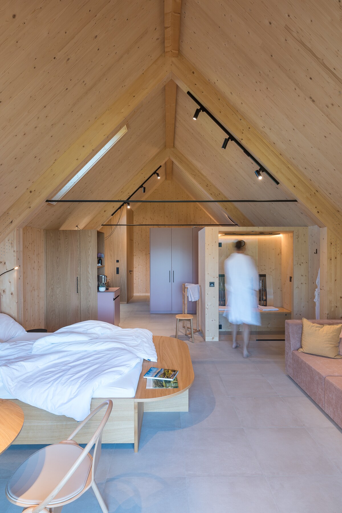 The spacious sleeping area features a large, unmade bed against a backdrop of natural wood paneling. A soft, neutral-toned sofa is present, along with a dining table crafted from light wood. Soft lighting and angled ceilings enhance the contemporary design of the room.