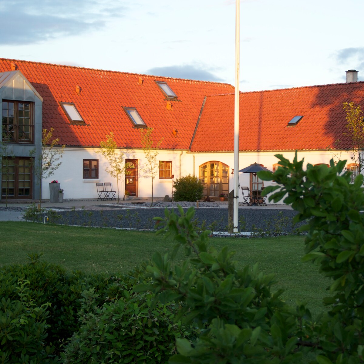 The exterior of the property features a charming building with a red-tiled roof and large windows. The landscaped garden includes seating arrangements, and a pathway leads to a shaded terrace under a parasol. Greenery and plants frame the scene, enhancing the sense of space.