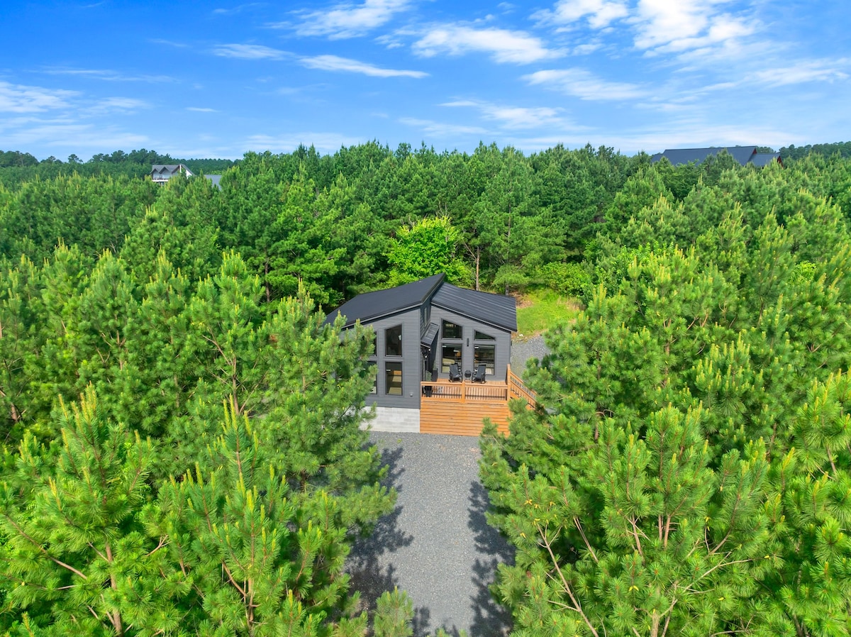 An aerial view captures the cabin nestled among tall, lush trees, highlighting its modern design with a black roof and large windows. The gravel path leading up to the cabin is flanked by greenery, creating a tranquil and private space in nature.