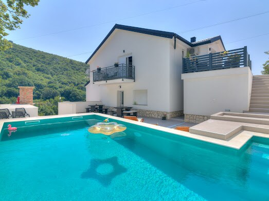 A modern two-story building features a spacious outdoor area with a turquoise pool. A sunny terrace with seating options is visible, and the surrounding landscape includes lush greenery. The upper balcony offers additional space for relaxation, enhancing the overall outdoor experience.