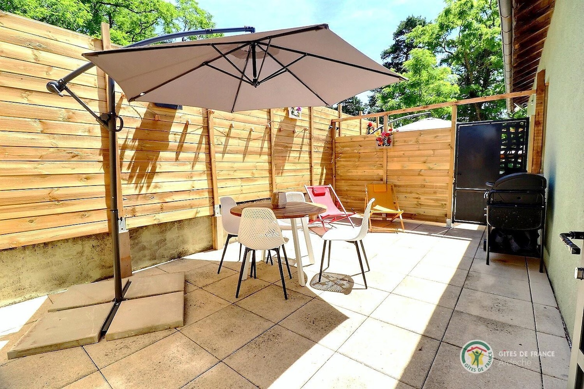 An outdoor terrace is equipped with a large umbrella providing shade over a round table surrounded by four chairs. A charcoal grill is visible, and a stylish lounge area includes two colorful chairs. Natural wooden fencing offers privacy to the space.