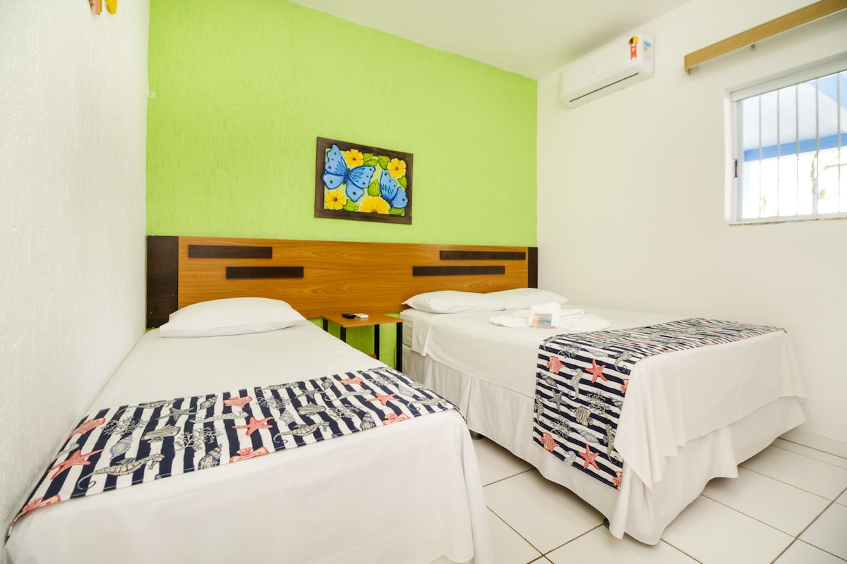 A bright guest room is featured, with a vibrant green accent wall. Two beds, one double and one single, are adorned with white linen and patterned throws. A colorful piece of wall art depicting butterflies is displayed above the beds. A window allows natural light to enter.