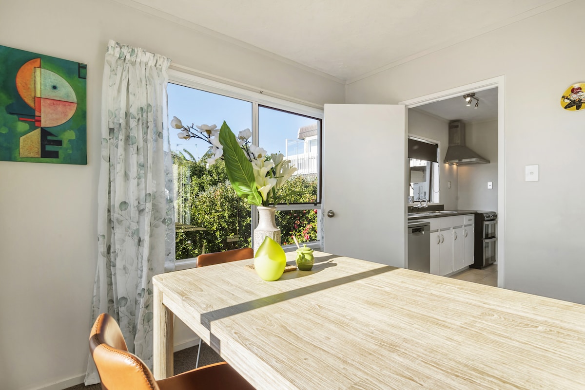 A dining area features a light wooden table complemented by tan chairs. A brightly colored artwork hangs on the wall beside a window, allowing natural light to illuminate the space. Lush greenery is visible outside, enhancing the inviting environment.