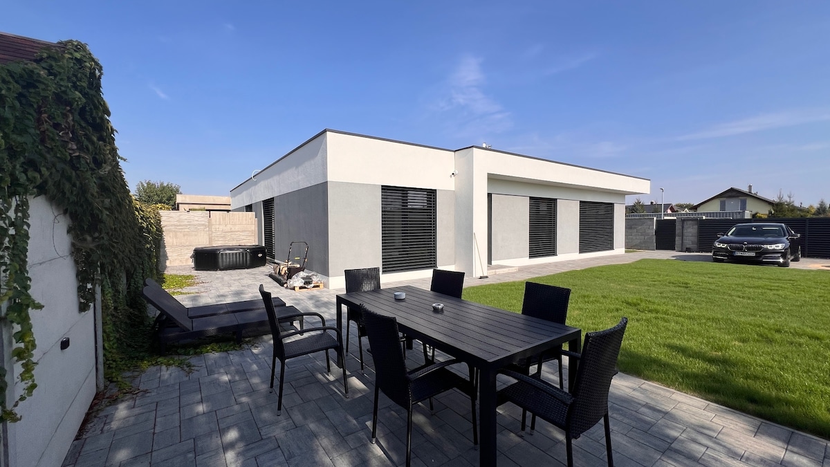 A modern house is presented from the garden, showcasing large windows and a sleek exterior. A spacious patio with a dark wooden table and black chairs is visible, surrounded by well-maintained grass and paving. A hot tub is positioned nearby, adding to the outdoor amenities.