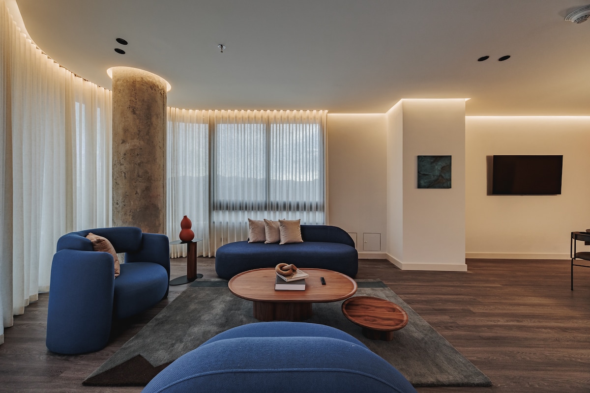 A spacious living area features rounded blue sofas that invite relaxation. A wooden coffee table sits centrally on a textured rug, complemented by sculptural decor. Natural light filters through sheer curtains, while soft ambient lighting enhances the warm, inviting space.