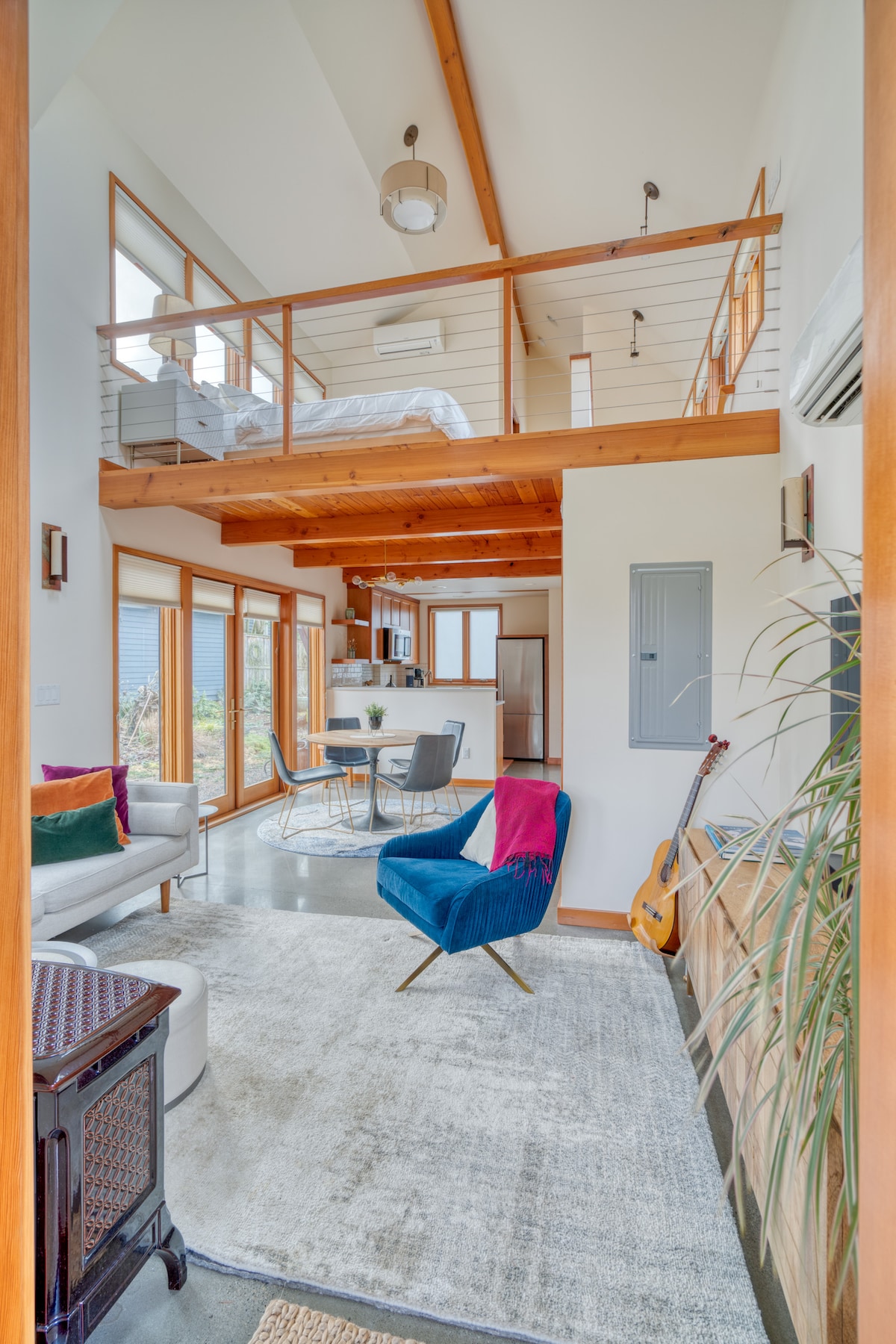 A bright and airy living area is highlighted by large windows that invite natural light. A dining table and chairs are visible, along with a cozy seating arrangement. An upper loft area can be seen, with a spacious bed partially in view, enhancing the open layout.