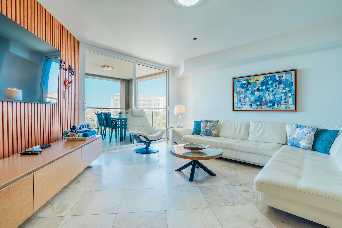 The living area features a spacious white sectional sofa, accented by decorative cushions. A wooden accent wall complements a sleek entertainment unit with a mounted TV. Natural light enters through large sliding glass doors that lead to the balcony. Additional seating is available with a modern chair and a round coffee table.