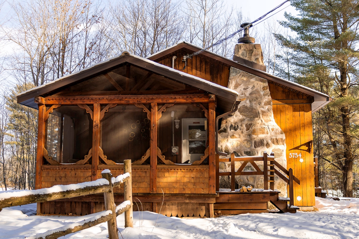 The exterior of the chalet showcases natural wood siding and a prominent stone chimney. A screened porch is featured on the front, complemented by wooden railings. Surrounding snow-covered ground is visible, along with tall trees creating a peaceful setting.