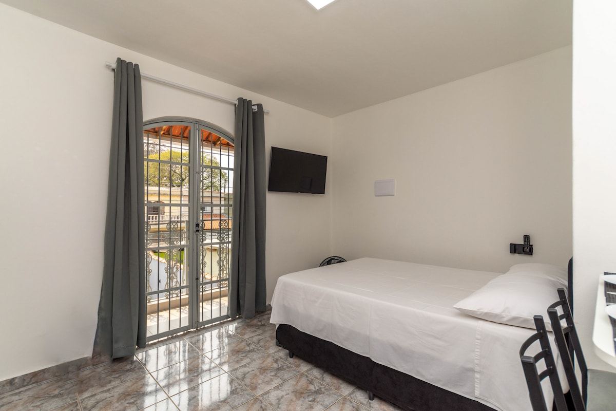 A bright room features a spacious bed dressed in crisp white linens. Natural light enters through a set of glass doors with iron bars, leading to an outdoor area. A wall-mounted television is positioned across from the bed, and dark curtains frame the window.