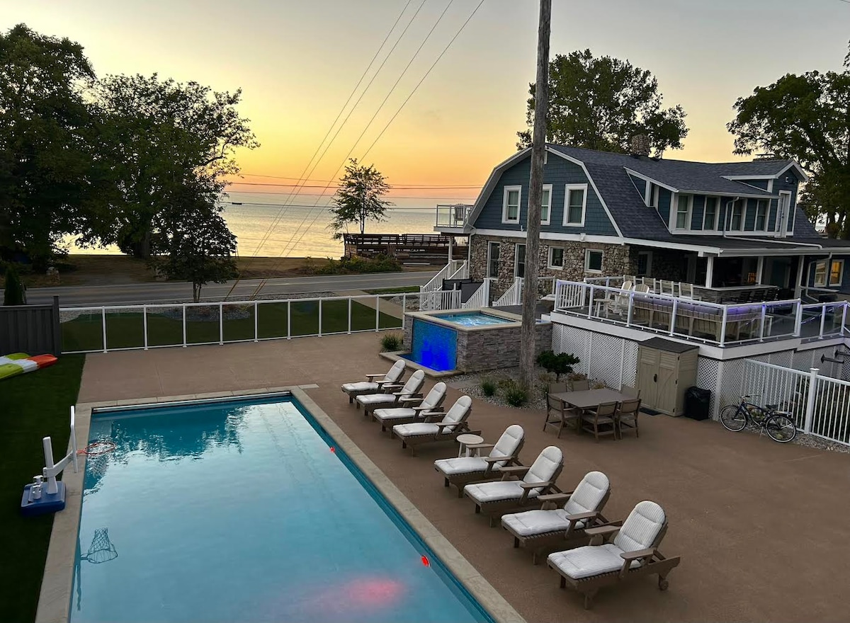 A scenic view features a large pool surrounded by lounge chairs and a dining table. The backdrop includes a multi-level house with outdoor seating and a sunset over the lake, offering a tranquil atmosphere. Tall trees line the property, enhancing the natural setting.