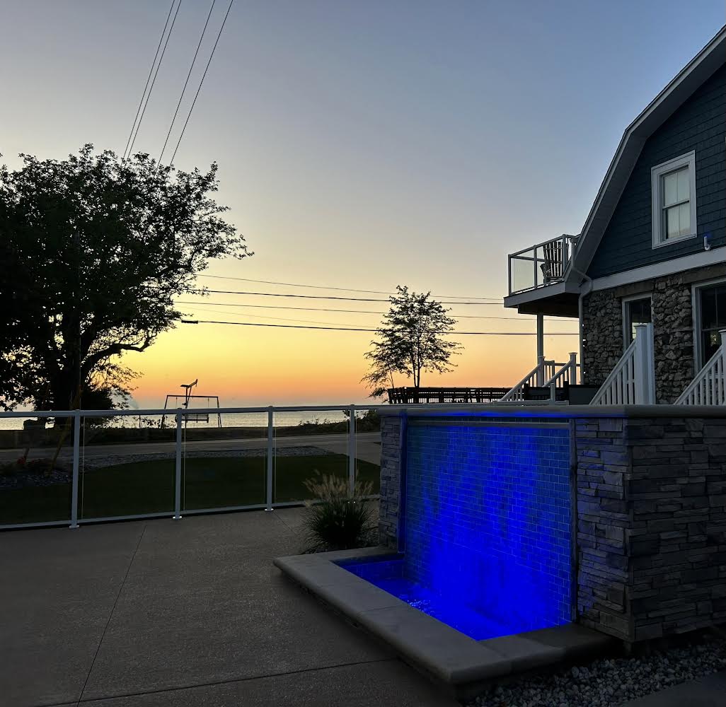 An outdoor space features a sleek water feature illuminated in blue, set against a serene sunset sky. The background includes a large tree and a building with a stone facade, while a deck is visible above, offering a view of the surrounding landscape.