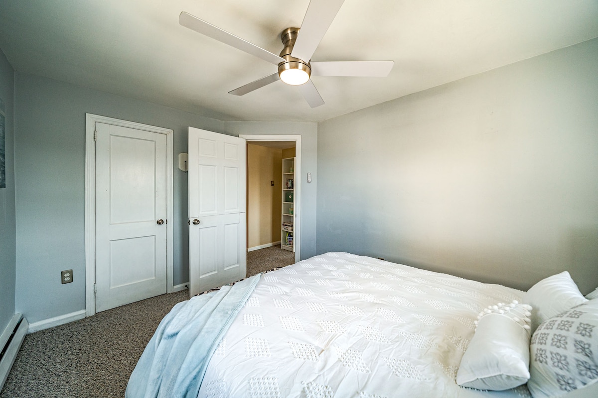 A well-lit bedroom features a spacious bed adorned with crisp white linens. A ceiling fan provides airflow, and light-colored walls contribute to a bright atmosphere. Two doors lead to additional areas, enhancing accessibility and convenience.