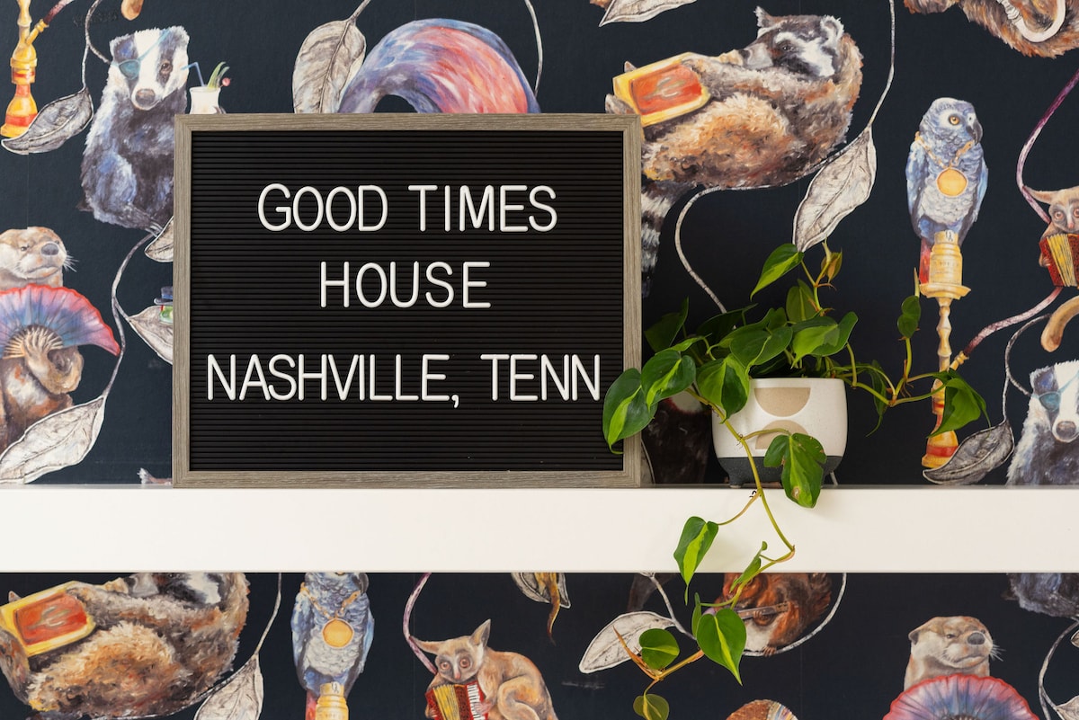 A detailed wall is adorned with a whimsical animal-themed wallpaper featuring various creatures. In the foreground, a letter board displays the text 'GOOD TIMES HOUSE NASHVILLE, TENN' surrounded by a green plant in a white ceramic planter.