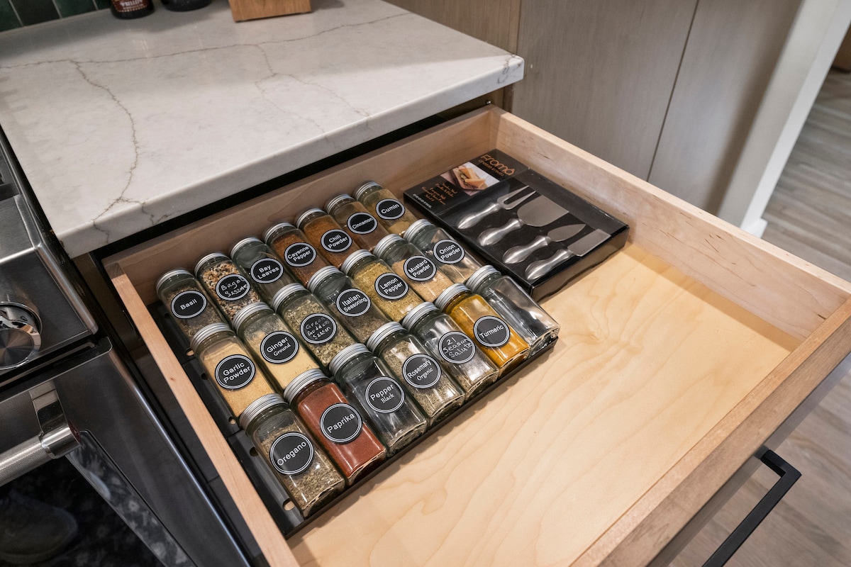 A kitchen drawer is organized with a selection of labeled spice jars arranged neatly on a tray. A set of measuring spoons is positioned beside the spices, all on a clean, lightwood base. The countertop is visible in the background, featuring a marble surface.