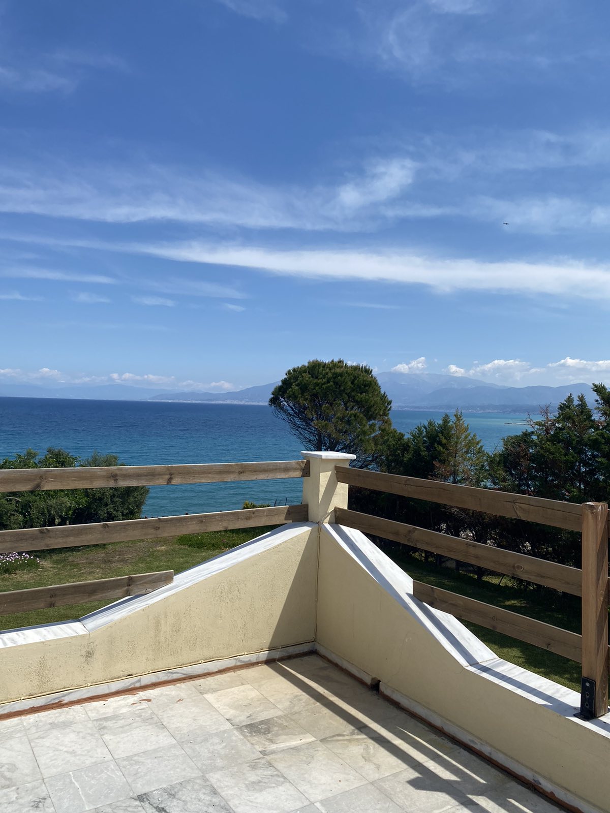 A spacious balcony overlooks the sea, framed by wooden railings. The view includes a clear sky, calm waters, and distant mountains, with greenery visible in the foreground. Soft light enhances the tranquil setting, creating an inviting atmosphere for relaxation.