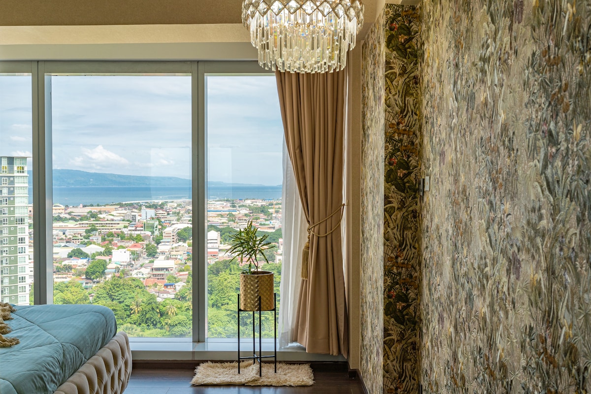 A spacious bedroom features a super king size bed, beautifully contrasted against floral luxury wallpaper. Natural light floods through large glass windows, offering expansive views of Davao City, with glimpses of green landscapes and distant mountains. A decorative chandelier hangs above, adding an elegant touch.