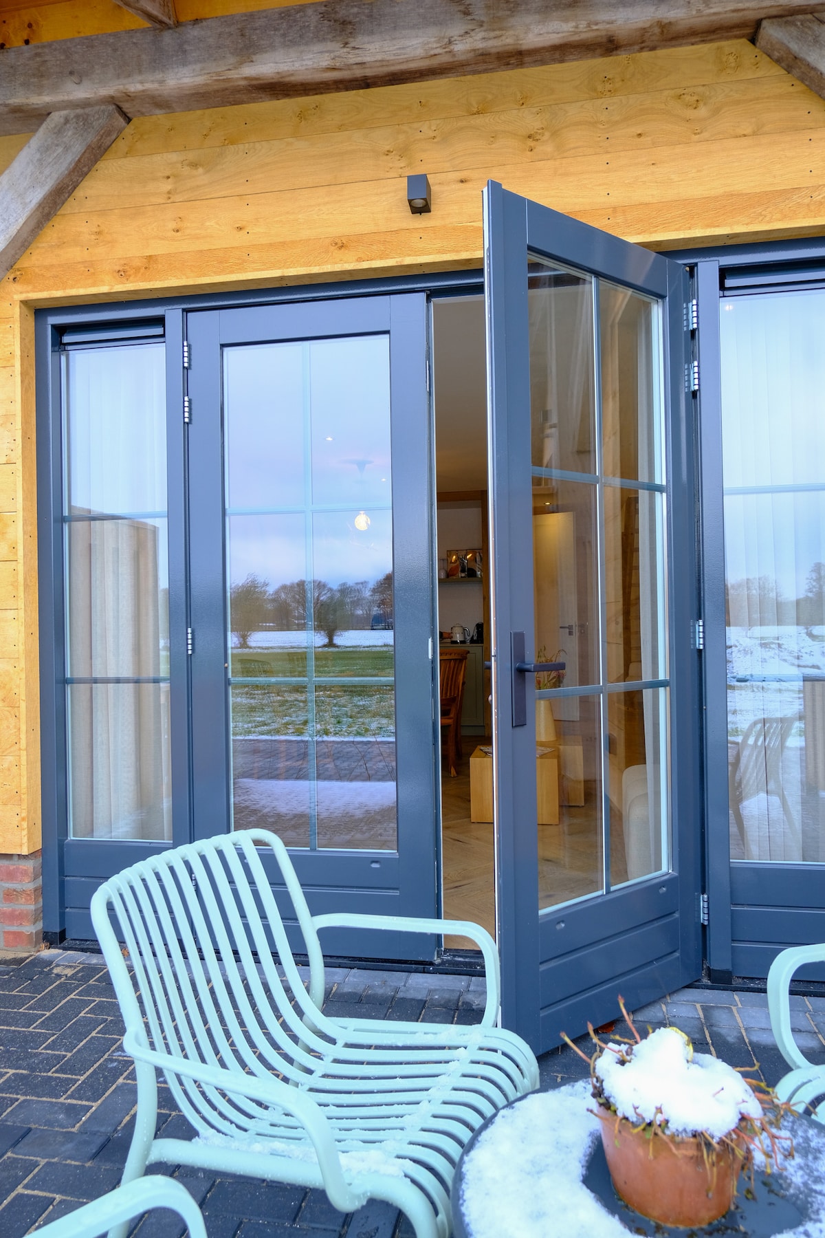 A pair of double glass doors opens to reveal a welcoming entrance, framed by a wooden structure. A light turquoise chair is positioned outside, alongside a potted plant topped with snow. The expansive landscape is visible beyond the doors, emphasizing the serene location.