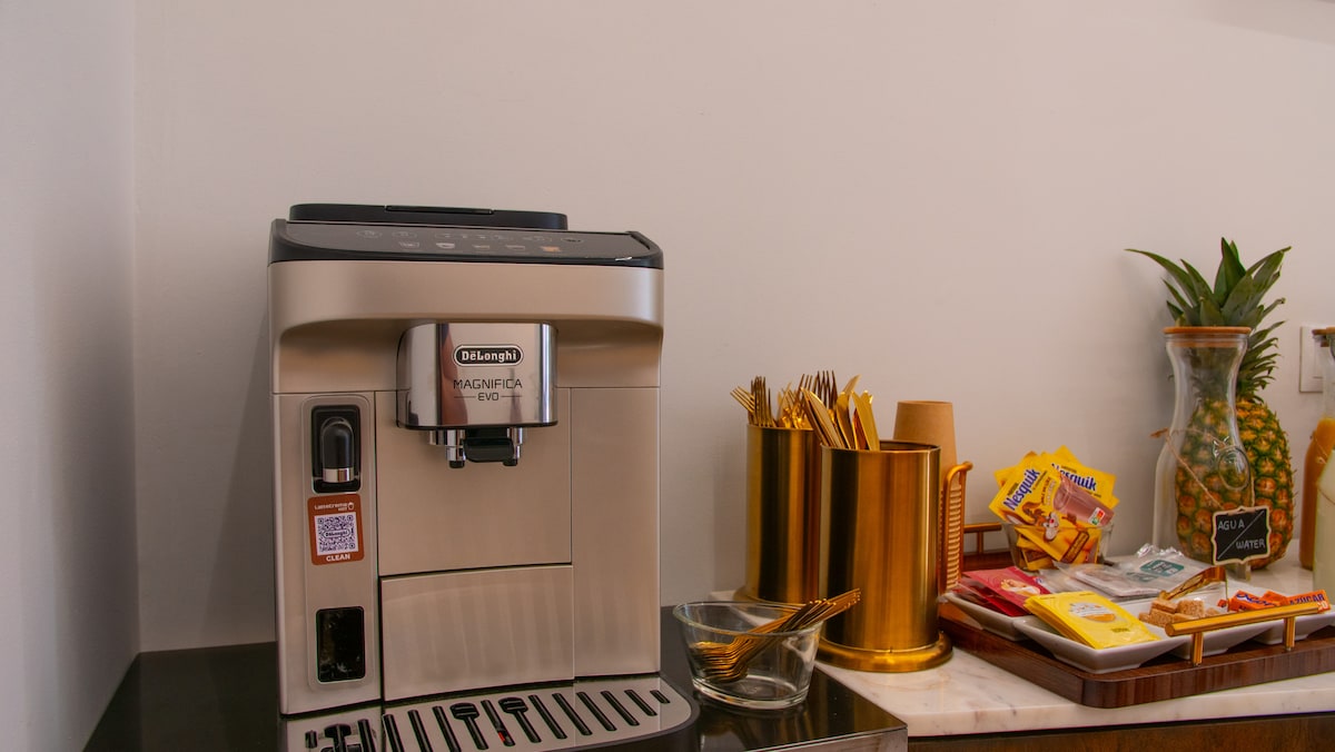 A modern coffee machine is positioned on a countertop alongside an array of utensils in a gold holder. Packaged snacks and a pineapple add a decorative touch to the service area, creating a welcoming setting for guests to enjoy refreshments.