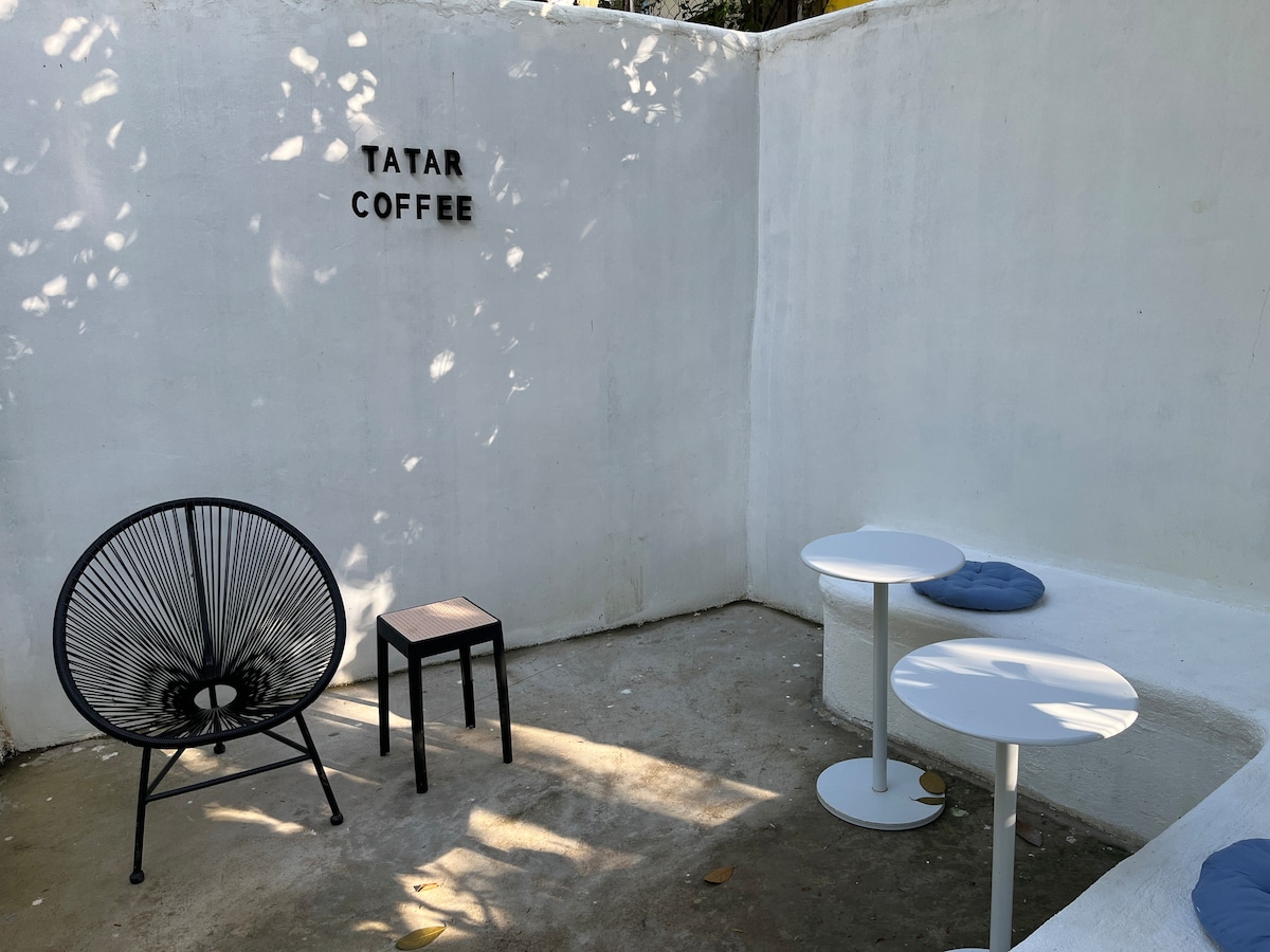 An outdoor seating area is shown with a black, round wicker chair and a small, dark wooden chair positioned near two white tables. The walls are painted white, and a sign reading 'TATAR COFFEE' is mounted on the wall, casting soft shadows in the daylight.