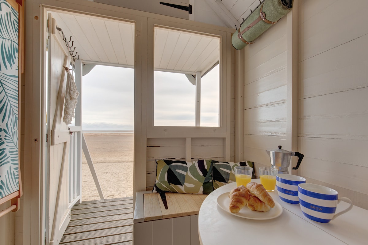 The interior of the beach hut features a cozy seating area with a decorative cushion. A simple table is set with a plate of croissants, a coffee maker, and a glass of orange juice. Large windows provide a view of the sandy beach.