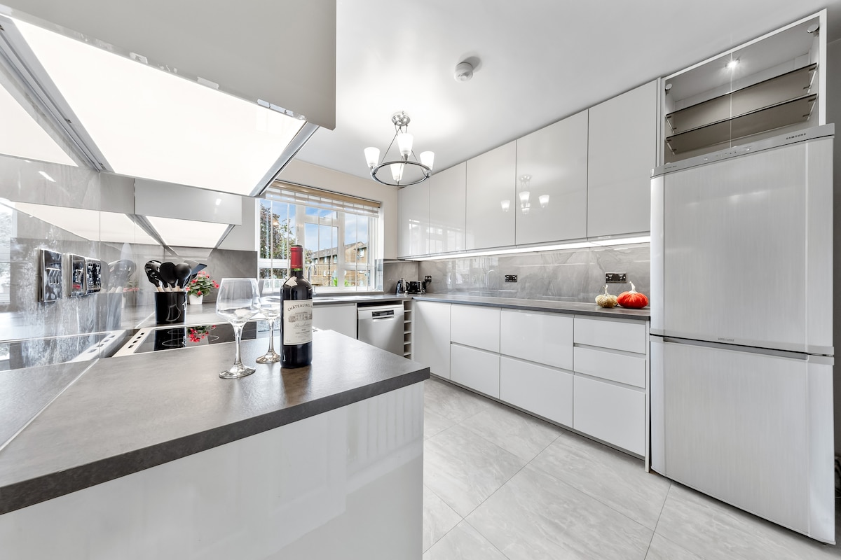 The modern kitchen features sleek white cabinetry and a spacious island with a dark countertop. Natural light enters through large windows, illuminating the area. A dining space can be seen, with a bottle of wine and garnishes on the counter, creating a stylish and functional environment.