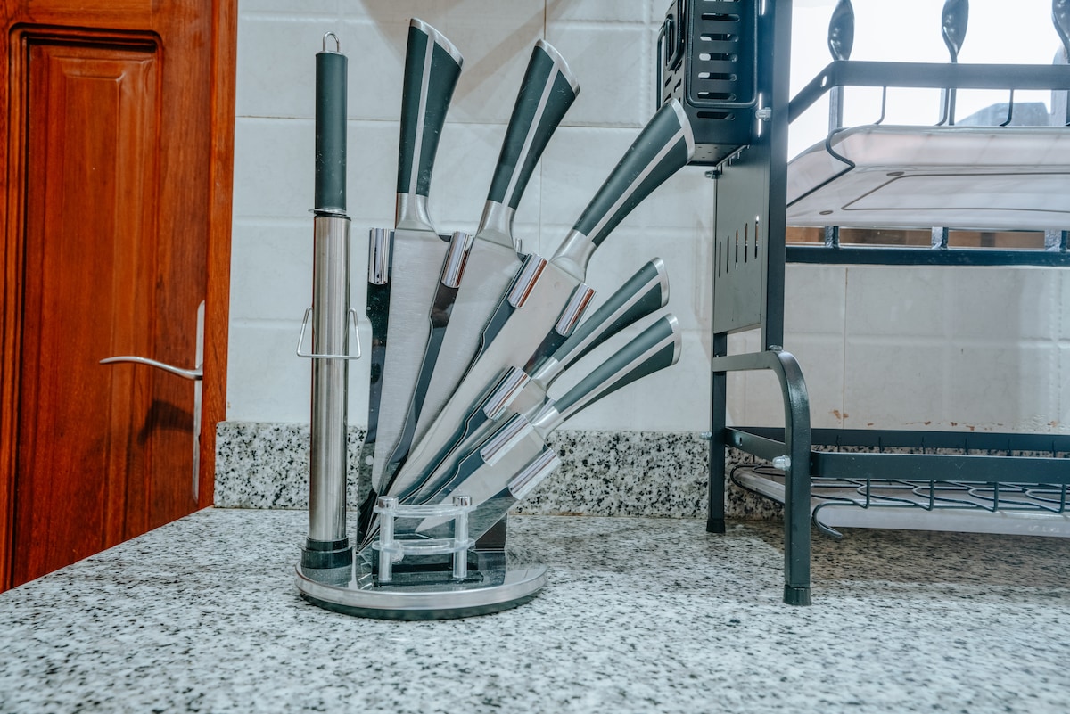 A knife set is organized on a granite countertop, featuring various sizes of knives arranged in a sleek stand. A wooden cabinet and a metal rack are visible in the background, complementing the kitchen's functionality.